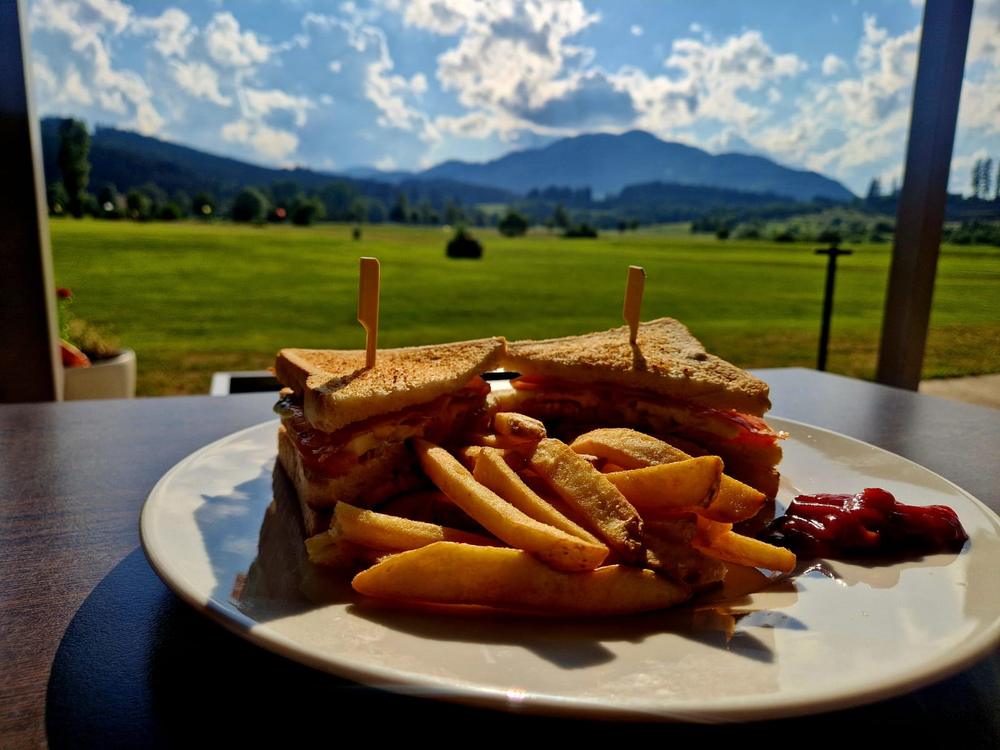 Club Sandwich mit Pommes am Tisch, dahinter die Wiesen und die Berge